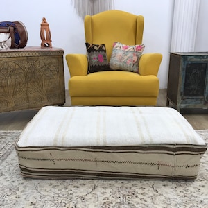 May include: A white and brown striped ottoman with a thick, woven fabric. The ottoman is sitting on a patterned rug in front of a yellow armchair and a wooden chest.