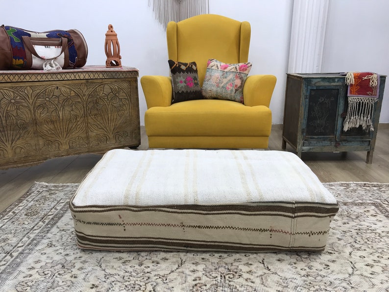 May include: A white and brown striped ottoman with a thick, woven fabric. The ottoman is sitting on a patterned rug in front of a yellow armchair and a wooden chest.