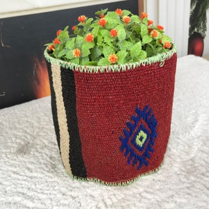 Puede incluir: Un macetero tejido rojo con un diseño geométrico en azul y verde. El macetero tiene un borde a rayas blanco y negro y está lleno de plantas verdes con pequeñas flores naranjas.