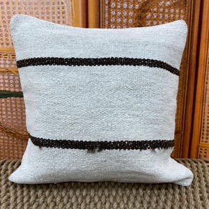 May include: A square, light blue pillow with two dark brown horizontal stripes. The pillow has a textured appearance and is likely made of woven fabric. The stripes are evenly spaced across the pillow's surface, adding a simple, yet stylish design.