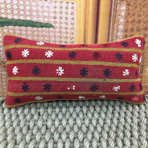 Puede incluir: Una almohada rectangular roja con un patrón de rayas. La almohada presenta rayas doradas horizontales y está decorada con diseños florales blancos y azul marino. La almohada está colocada sobre una superficie tejida.