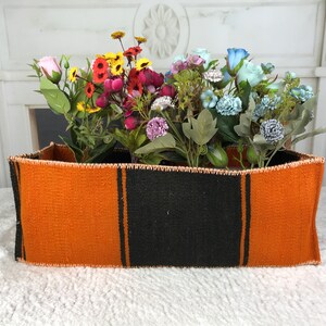 Può includere: Un cesto rettangolare intrecciato di colore arancione e nero con una varietà di fiori artificiali colorati all'interno.