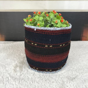 Op de afbeelding: Een geweven plantenbak met een blauw, zwart en rood gestreept patroon. De plantenbak heeft een witte rand aan de bovenkant en is gevuld met een groene plant met kleine oranje bloemen.