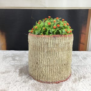 Pode incluir: Um vaso cilíndrico de juta trançada com uma superfície bege texturizada e bordas costuradas com fio vermelho na parte superior e inferior. O vaso contém uma pequena planta artificial com folhas verdes e pequenas flores cor de laranja.