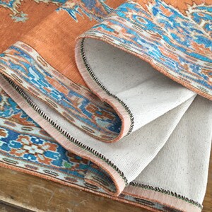 May include: A close-up of a folded orange and blue patterned rug with a white backing. The rug has a black stitched border.