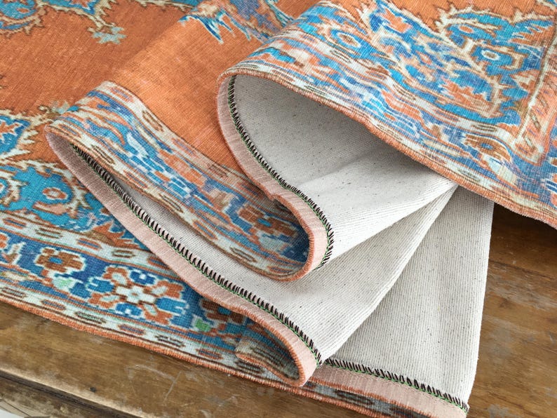May include: A close-up of a folded orange and blue patterned rug with a white backing. The rug has a black stitched border.