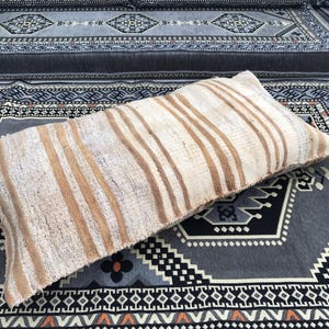 May include: A long, rectangular, off-white pillow with brown stripes. The pillow is lying on a gray and black patterned rug.