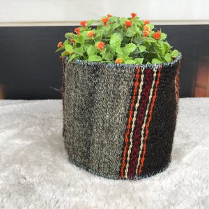 Op de afbeelding: Een cilindervormige mand van geweven stof met een gestructureerd grijs en zwart patroon met verticale strepen in rood, oranje en crème. De mand bevat een kleine kunstplant met groene bladeren en kleine oranje bloemetjes.