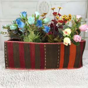 Puede incluir: Un macetero rectangular tejido a rayas con un esquema de color rosa, marrón y naranja. El macetero está lleno de una variedad de flores artificiales, incluyendo rosas azules, rosas y amarillas.