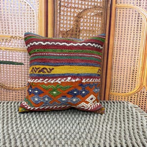 May include: Square decorative pillow with a vibrant, geometric pattern. The pillow features a mix of colors including red, green, yellow, and blue. The design includes diamond and zigzag patterns, creating a textured appearance. The pillow is placed on a gray, woven surface.