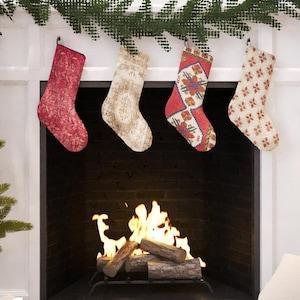 Puede incluir: Cuatro calcetines navideños decorativos, rojos y crema, cuelgan sobre una chimenea con fuego. Los calcetines están suspendidos de una repisa blanca, con una guirnalda de vegetación encima.