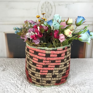 May include: A woven basket with a pink and brown geometric pattern holds a bouquet of pink, blue, and yellow flowers.