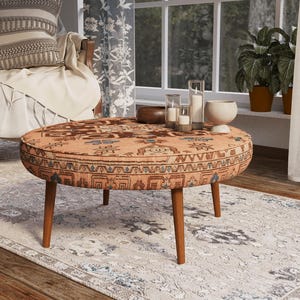 May include: A round coffee table featuring a patterned fabric top in shades of orange, brown, and blue with a geometric and floral design. The table is supported by four tapered wooden legs and has a low-profile, circular silhouette.