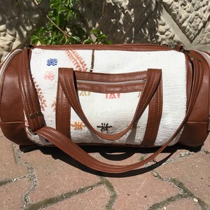 Puede incluir: Una bolsa de viaje blanca y marr&oacute;n con ribete de cuero y cierre de cremallera. La bolsa tiene un dise&ntilde;o bordado de colores.