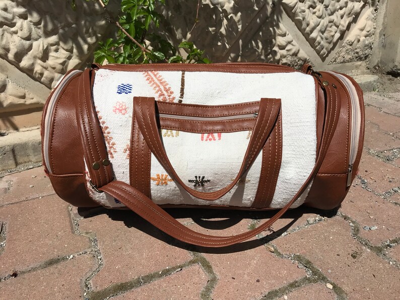Puede incluir: Una bolsa de viaje blanca y marr&oacute;n con ribete de cuero y cierre de cremallera. La bolsa tiene un dise&ntilde;o bordado de colores.