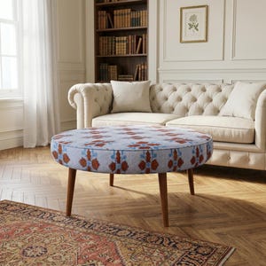 May include: A round ottoman featuring a light gray fabric upholstery with a repeating floral pattern in shades of rust orange and blue. The piece is supported by four tapered wooden legs in a medium brown finish, providing a stable base.