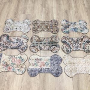May include: Nine bone-shaped rugs with distressed patterns in various colors, including blue, beige, and brown. The rugs are arranged on a light wood floor, showcasing their unique designs and textures. These rugs are ideal for pet owners.