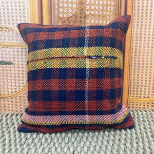 May include: Square pillow with a plaid pattern in red, navy blue, yellow, and pink. The pillow has a zipper closure and is made of a woven fabric. The pillow is approximately 16 inches square.