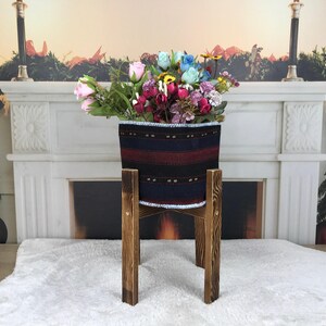 Puede incluir: Una maceta cilíndrica de tela con rayas horizontales en negro, rojo oscuro y azul, sostenida por un soporte de madera de cuatro patas con un acabado teñido oscuro. La maceta contiene un arreglo de flores artificiales.