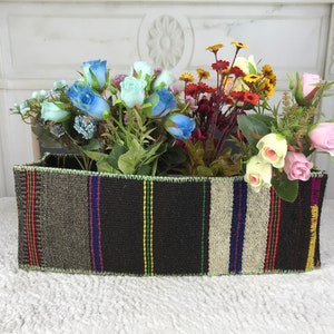 May include: A woven rectangular planter with a colorful striped pattern holds a variety of artificial flowers, including blue, pink, and yellow roses.