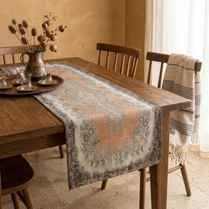 Può includere: Runner da tavolo rettangolare con un intricato motivo floreale nei toni del pesca, grigio e verde, bordato da un bordo marrone scuro. Posizionato su un tavolo di legno, con una brocca di rame e bicchieri. Lunghezza circa 150 cm.