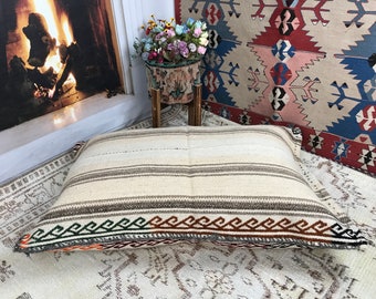 May include: A white and brown striped floor cushion with a woven border in shades of green, orange, and brown. The cushion is sitting on a white rug.