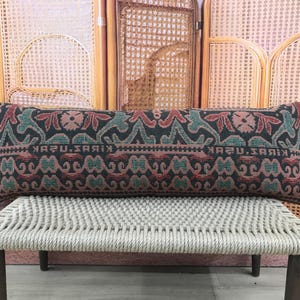 May include: Rectangular decorative pillow with a patterned design in shades of black, teal, and red. The pillow is resting on a light-colored woven bench. The pillow's pattern includes floral and geometric elements, with text visible.