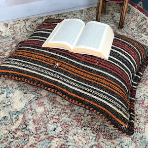 May include: A striped floor cushion with orange, black, and white stripes. The cushion is made of woven fabric and has a fringed edge. A book is resting on top of the cushion.