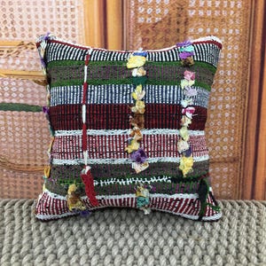Peut inclure: Un coussin carré décoratif avec un motif tissé. Le coussin présente des rayures horizontales rouges, blanches, vertes et noires. Des glands colorés et des accents verticaux rouges et blancs ajoutent des détails.