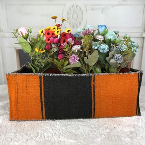 Puede incluir: Una cesta de almacenamiento rectangular con textura tejida y rayas verticales alternas en naranja brillante y negro. La cesta está llena de un surtido de flores artificiales en tonos rosa, amarillo, rojo, morado y azul claro.