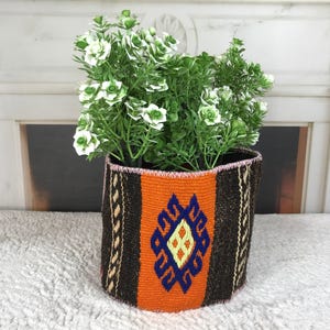 Könnte beinhalten: Ein zylindrischer geflochtener Korb mit einem leuchtend orangefarbenen Mittelteil mit blau-gelbem geometrischem Rautenmuster, flankiert von dunkelbraunen, strukturierten Seiten mit cremefarbenen vertikalen Streifen, gefüllt mit einer grünen Kunstpflanze.