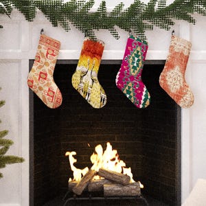 Könnte beinhalten: Vier gemusterte Weihnachtsstrümpfe hängen über einem Kamin mit einem brennenden Feuer. Die Strümpfe weisen verschiedene Designs in Rot, Gelb, Rosa und Orange auf. Links ist ein Weihnachtsbaum mit Dekorationen.