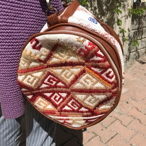 Puede incluir: Un bolso redondo tejido de color blanco y marr&oacute;n con un patr&oacute;n geom&eacute;trico. El bolso tiene un ribete de cuero marr&oacute;n y una cremallera.