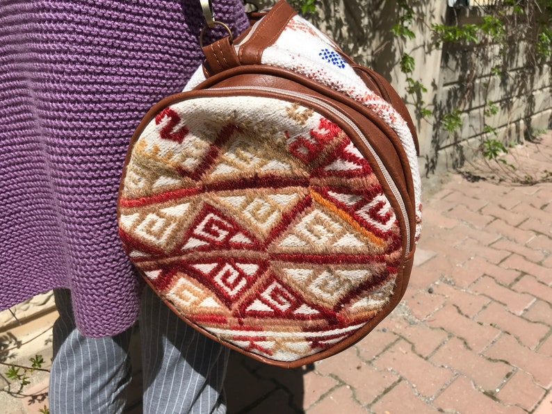 Puede incluir: Un bolso redondo tejido de color blanco y marr&oacute;n con un patr&oacute;n geom&eacute;trico. El bolso tiene un ribete de cuero marr&oacute;n y una cremallera.