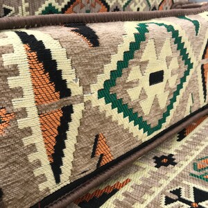 May include: A close-up of a brown and beige patterned throw pillow with a dark brown border. The pillow has a geometric design with green, orange, and black accents.