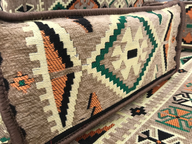 May include: A close-up of a brown and beige patterned throw pillow with a dark brown border. The pillow has a geometric design with green, orange, and black accents.
