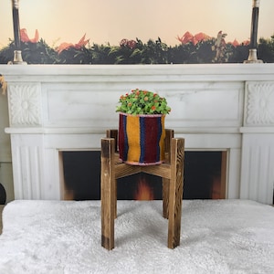 以下が含まれることがあります： カラフルなストライプのプランターに入った鉢植えの植物が置かれた木製プランタースタンド。プランタースタンドは、緑色のガーランドが飾られた白い暖炉の前にある。