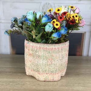 Puede incluir: Un macetero de mimbre tejido con un patrón rosa, verde y blanco, que contiene un ramo de flores azules y amarillas.