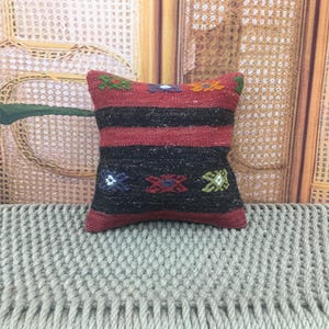 May include: A small, rectangular pillow with a woven design. The pillow features alternating bands of red and black, with colorful geometric patterns. The pillow is placed on a light gray, textured surface.