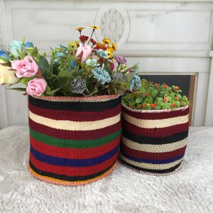 May include: Two cylindrical woven baskets featuring horizontal stripes in black, cream, maroon, green, and blue. The larger basket contains a bouquet of artificial pink, yellow, and blue flowers, while the smaller basket holds green foliage.