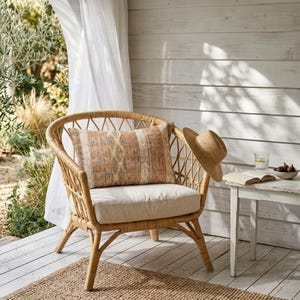 May include: A wicker chair with a beige cushion and a patterned pillow. A straw hat hangs on the chair's back. A small white table holds a glass of water, a book, and a bowl of figs. The chair sits on a wooden deck with a woven rug.