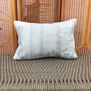 May include: Off-white rectangular cushion with vertical stripes and patches. It rests on a woven surface. The background shows a wooden structure with a woven pattern. The cushion has a rustic, vintage appearance.