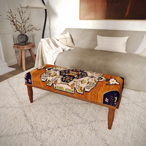 Puede incluir: Banco rectangular con una parte superior estampada en tonos naranja, azul marino y crema. El banco tiene cuatro patas de madera y está colocado sobre una alfombra blanca y peluda. El diseño de la parte superior del banco presenta motivos geométricos y florales.