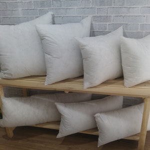 May include: A set of eight white pillow inserts of various sizes, arranged on a wooden shelf against a white brick wall.