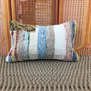 May include: Rectangular decorative pillow with a patchwork design. The pillow features a mix of colors including blue, white, and various earth tones. The pillow is placed on a textured, woven surface, suggesting a rustic or bohemian style.