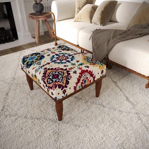 May include: A square ottoman featuring a cream-colored fabric upholstery with a vibrant, multi-colored floral and geometric pattern in shades of red, blue, yellow, and green. The piece is supported by four dark brown wooden legs with a tapered design.