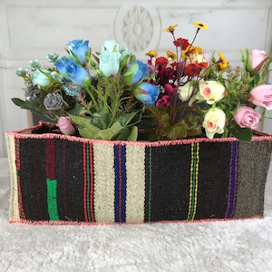 Può includere: Un cesto rettangolare intrecciato pieno di fiori artificiali colorati. Il cesto ha uno sfondo scuro con strisce verticali in vari colori, tra cui rosso, verde e blu. I fiori includono rose blu, rosa e gialle.