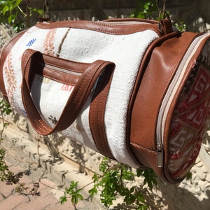 Puede incluir: Una bolsa de viaje cil&iacute;ndrica blanca y marr&oacute;n con cierre de cremallera y un dise&ntilde;o estampado. La bolsa tiene dos asas de cuero marr&oacute;n.