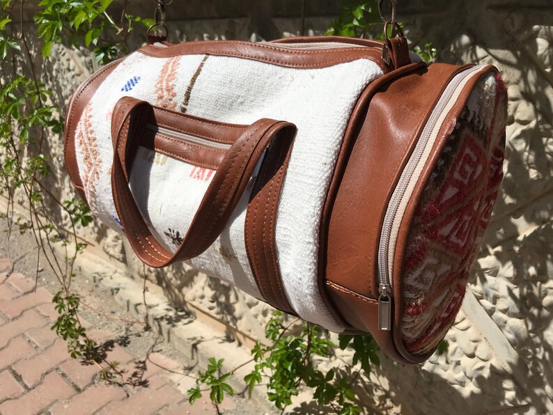 Puede incluir: Una bolsa de viaje cil&iacute;ndrica blanca y marr&oacute;n con cierre de cremallera y un dise&ntilde;o estampado. La bolsa tiene dos asas de cuero marr&oacute;n.