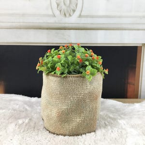 May include: A small, cylindrical planter wrapped in a textured, light brown burlap fabric. The planter contains a dense cluster of artificial green foliage accented with numerous tiny, bright orange flower buds distributed throughout the plant.